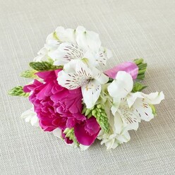 A small but striking floral arrangement lies horizontally across a pale beige, linen-textured cloth, its subtle weave echoing the relaxed, townhouse charm of Canonbury's quiet terraces. The eye is immediately drawn to a sumptuous central bloom in vivid fuchsia, most likely a peony or garden rose, with layers upon layers of ruffled petals that curl and fold into one another, creating a plush, cushion-like centre. This bold pink heart is softened and refined by several white alstroemeria blooms cradling it on either side, their slender trumpet-shaped petals gently arching outward. Inside each white petal, fine maroon and burgundy lines radiate from the throat, forming intricate patterns that feel almost like handwritten script across the flowers' surfaces. Peeking out between the main blooms are delicate, elongated buds and spiky florets in fresh spring green, giving the piece a sense of movement and adding a touch of untamed garden energy. The stems are carefully aligned and bound together with a smooth satin ribbon in bright magenta, its sheen catching the light and reinforcing the romantic, modern palette. The lighting is soft and natural, as if filtering through a Canonbury bay window on an overcast afternoon, enhancing the gentle shadows and highlights that bring depth to every petal. The arrangement feels designed as a boutonniere, corsage, or intimate posy for a wedding or prom-something you might wear to a ceremony at a local church near Canonbury or a celebration in a nearby townhouse garden. It radiates warmth, closeness, and the slightly fluttery excitement that comes just before you step into a special moment.