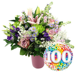 Set against a crisp white background, this lavish centenary bouquet feels like a miniature celebration captured in flowers, the sort of thoughtful gift you might send to a long-time Canonbury neighbour who has seen generations grow up along nearby St Paul's Road. A matte, ribbed pink vase anchors the design, its soft blush tone echoing the pastel palette of the blooms. Rising from it, tall pale pink Oriental lilies create the main focal point, some fully unfurled with sweeping petals and pollen-tipped stamens, others still wrapped in smooth buds that suggest quiet anticipation. Around them, layers of soft pink and dusky mauve roses form a romantic heart, their gently curling petals giving the impression of time and memories unfolding. Deep purple lisianthus stems thread through the arrangement like ribbons of satin, complemented by dainty lavender asters whose tiny golden centres sparkle against their cool-toned petals. Bright lime green button chrysanthemums add playful, rounded notes of texture, while wisps of white baby's breath drift between stems like light Canonbury morning mist. Lush green foliage, in varied shapes from broad leaves to slender stems, adds structure and contrast, making the pastel shades glow even more intensely. To the lower right of the flowers, a gleaming round foil balloon introduces a burst of colour and fun. It is fully covered in multi-coloured confetti dots in shades of red, orange, yellow, green, and blue, with the number "100" standing out boldly in large white numerals with a subtle shadow, signalling a proud century. The overall composition feels festive yet elegant, perfectly suited for marking a 100th birthday, a centennial wedding anniversary, or any remarkable lifetime achievement in the Canonbury community.