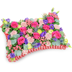 Seen from a slightly raised oblique viewpoint on a pristine white surface, this pillow-style memorial arrangement is tightly packed and meticulously balanced, every bloom placed to create a plush, continuous surface. The composition is anchored by numerous hot-pink roses, their velvety petals opening to reveal warm, saturated centres that glow against the neutral backdrop. Interlaced among them are soft, pale-pink carnations that contribute a ruffled, tactile layer, and deep purple lisianthus whose bell-like forms draw the eye inward. Cheerful lemon-yellow ranunculus provide bright, sunlit notes across the cushion, while sprays of silver-blue eryngium introduce spiky, sculptural contrast and a cooler colour temperature. Lush greenery - notably rounded eucalyptus leaves with their silvery bloom and other mid-green foliage - knits the flowers together, offering both structure and a subtle eucalyptus freshness. The perimeter is finished with a ruffled red satin ribbon that frames the pillow with a refined, satin sheen and a slight pleat that catches the light. The studio lighting is soft and even, highlighting petal texture and the careful layering that gives the pillow its supportive, pillowy profile. Intended as a sympathetic floral tribute, the palette leans toward a celebration of cherished memories: vivid fuchsia and violet offset by gentle blush and lemon tones to comfort rather than mourn. As a local Islington florist might explain, this kind of cushion is often chosen for services around Essex Road or New River Walk where families seek a bright, consoling expression of remembrance.