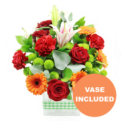 In this detailed product image, a lush, densely packed bouquet seems to burst from a sleek rectangular glass vase, every bloom carefully positioned to draw the eye. At the centre, several deep red roses, with tightly furled, velvety petals, offer a classic focal point, their rich colour echoing the brick tones of Canonbury's terraces. Interwoven with them are red-orange carnations, the ruffled edges of their petals creating a sense of movement and layered texture, as though the bouquet is in gentle motion. Radiant orange gerbera daisies with dark brown or almost black centres sit slightly forward, their flat, symmetrical faces bringing a joyful, contemporary note that would brighten any table off Upper Street. Towering above, white Oriental lilies rise on strong stems; some are fully open, revealing pink-tinged throats and prominent stamens, while others remain in tight buds, adding a vertical rhythm and promise of continued blooming. Between the larger flowers, small, round lime-green pompom chrysanthemums nestle like bright beads, their compact, cushion-like forms offering a fresh, zesty contrast. Rich green foliage, including broad, glossy leaves and sprigs of silvery eucalyptus, surrounds and supports the blooms, creating layers of depth and a natural, garden-fresh feel. The stems are clearly visible through the transparent sides of the modern rectangular vase, which is wrapped near its base with a crisp light green and white gingham ribbon, giving a hint of country charm amid the city setting. In the bottom right corner of the image, a bold circular orange overlay displays the words "VASE INCLUDED" in clean white capitals, clearly indicating that this chic container is included, making the arrangement an effortless, ready-to-enjoy gift for birthdays, congratulations, or welcoming a new neighbour in Canonbury.