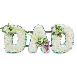 The image presents a lovingly crafted funeral tribute spelling DAD in sturdy three-dimensional letters, a design that would feel at home at a quiet Canonbury service or a farewell near Highbury Corner. Each letter is completely carpeted in crisp white chrysanthemums, their tight, button-like blooms placed so closely that no gaps appear, creating a plush, almost velvety texture. They catch the light softly, giving the tribute a calm, luminous quality against the plain white background. A rich emerald green satin ribbon, carefully pleated and ruffled, outlines every curve and corner of the letters, bringing definition and a touch of natural colour reminiscent of the tree-lined streets around Canonbury Square. Behind the flowers, the dark horizontal lines of the support frame peep through just enough to show how the structure stands upright as one united piece.   

Thoughtful accent flowers break the white expanse in just the right places. At the upper left of the first D, a small spray of white freesias and glossy green leaves emerges, their slender buds and open florets creating a sense of gentle movement. Two elegant green stems sweep diagonally from this first D across towards the centre of the A, guiding the eye to a striking focal arrangement. There, two deep purple calla lilies nestle among more white freesias and foliage, their rich, sombre hue adding depth and a dignified contrast. On the final D, a more compact cluster of freesia and greenery mirrors the first, completing the visual balance.   

This DAD tribute conveys quiet strength, gratitude, and sorrow, perfectly suited for a father's funeral, wake, or memorial gathering around Canonbury, whether placed beside a coffin, at a graveside, or near a display of family photographs.