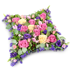 Set against a clean white background, this cushion-shaped funeral tribute appears like a soft, floral cloud, its edges gently rounded and inviting to the eye. The entire surface is tightly packed with roses and seasonal blooms, creating a rich tapestry of colour in shades of vivid fuchsia, warm pastel pink, and buttery cream. Many of the roses are fully open, revealing delicate spirals of petals that speak of love, gratitude, and cherished memories. Threaded gracefully through the roses are deep purple lisianthus, their velvety, ruffled flowers introducing a darker, contemplative note that lends the design a quiet nobility. Tiny lavender-toned filler flowers, such as statice or limonium, are scattered throughout like whispers of heather on a Hampstead Heath breeze drifting down towards Canonbury, softening the transitions between the larger blooms. Supporting this abundance is a lush bed of foliage: silvery eucalyptus leaves curve gently around the flowers, contrasting with darker green, fern-like fronds that build depth and texture. The resulting cushion looks both structured and tender, echoing the calm of Canonbury's garden squares and tree-lined streets. This arrangement is clearly intended as a sympathy or funeral tribute, perhaps resting at the front of a chapel or crematorium, where light falls quietly across each petal. It conveys a sense of serenity and thoughtful farewell, the sort of carefully considered piece families in Canonbury might choose when they want their final gesture to be both elegant and deeply heartfelt.