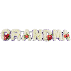 A horizontal tribute of floral lettering forms the word GRANDMA in large, clearly legible capitals, each one generously covered in snowy white spray chrysanthemums that create a soft, cushion-like surface. The flowers are so closely packed that almost no gaps in the petals can be seen, giving the impression of sculpted clouds of white. Around the outer edges of each letter, a narrow border of delicate pale purple ribbon or trim peeks through, adding depth and a gentle hint of colour that frames the name with calm refinement. The design is supported by a low, dark green frame that runs quietly behind the letters, giving the display enough height and structure to sit proudly at the front of a funeral service or beside a casket. Carefully placed accents of vivid red flowers bring emotional warmth to the tribute: the G is embellished with a cluster of deep red roses and a distinctive crimson-and-white lily or calla-shaped bloom; the first A holds a single, polished, heart-shaped anthurium; the M carries both red roses and another anthurium; and the last A is marked by a final red anthurium, balancing the composition. The contrast between the white base and the red focal flowers creates a moving symbol of enduring love, like cherished memories shining within grief. It is easy to imagine this piece crafted by Canonbury florists for a family in nearby Islington or De Beauvoir, then delivered with care to a local church, a simple yet powerful way to say goodbye to a dear grandmother.