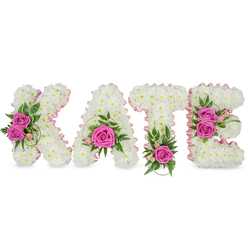 A carefully personalised floral name tribute from Canonbury Flowers spells KATE in large, sculpted letters, each formed from hundreds of immaculate white chrysanthemums that create a plush, seamless surface resembling freshly fallen snow. Soft pale-pink satin ribbon follows the inner contours of each character, lending a gentle, refined outline that subtly frames the bright floral typography. Hot-pink roses punctuate the white base as deliberate focal points: two open roses grace the left side of the K, a single bold bloom sits within the lower curve of the A, the stem of the T is marked by a vivid rose at its base, and the right arm of the E is warmed by a pair of deep pink roses. Mixed greenery - spiral grass, ruscus and variegated foliage - nestles beneath and around the roses, while delicate pale-pink spray blooms and small alstroemeria-style buds add a soft, garden-like texture. Displayed freestanding on a bright white surface, the letters cast gentle shadows that enhance the arrangement's depth and the florist's careful, compact packing of each chrysanthemum. The overall effect is luminous in cool daylight, the whites glowing and the pinks offering a compassionate, human touch. With its quiet dignity and handcrafted detail, this tribute is often chosen for funerals, memorials or celebrations of life in Canonbury, Islington and nearby Upper Street - available for sensitive local delivery when families prefer a personalised, respectful floral expression.