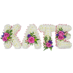 A carefully composed memorial tribute spelling KATE in four upright, three-dimensional letters, each formed from a dense mat of immaculate white chrysanthemums that give the letters a plush, tactile presence. Pale pink satin ribbon is used to outline each character, tracing both outer edges and inner counters with a soft, luminous line that lends tenderness to the overall silhouette. Each letter bears strategic accent clusters: deep fuchsia-pink roses and smaller, sweet pastel pink spray roses are paired with notes of deep purple or blue flowers - likely statice or small lisianthus - and arranged with mixed foliage including delicate fern fronds and rounded eucalyptus leaves. The floral placement is deliberate - a strong cluster on the diagonal stroke of the K, one near the lower-right curve of the A, a compact grouping at the base of the T, and two carefully balanced clusters on the E's centre and lower bars - creating rhythm and a comforting visual cadence. The scents are low and layered: the warm rose perfume tempered by the subtle, fresh green of eucalyptus. Lit softly against a white background, the contrasts of pure white, vivid fuchsia, gentle pink and dusky purple feel both respectful and warm. Crafted with the attention to detail you'd expect from a Canonbury florist serving the local community from Canonbury Square to Essex Road, this bespoke name tribute is designed for funeral services, quiet wakes or celebration-of-life ceremonies - a deeply personal way to honour a loved one.