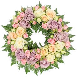 Centered on a clean white background, a lush circular funeral wreath appears almost like a soft ring of light, entirely covered with roses in gentle pastel tones and framed by deep green ferns. The flowers are packed closely together so that the eye reads them as a seamless tapestry of petals rather than individual stems. Dominating the design are fully open roses in creamy white and pale ivory, their broad, layered blooms suggesting tenderness and grace. Scattered among them are clusters of small blush pink spray roses, some still in tight bud and others half-unfurled, adding texture and a sense of life continuing onward. Here and there, petals in pale peach and buttery yellow introduce a mellow warmth, while occasional dusty lavender and muted mauve roses bring a vintage, romantic note that feels especially poignant in the context of loss. Around and between the blooms, slender fronds of dark green fern emerge in arcs and curves, giving the wreath a natural, breathing quality, as if it had just been gathered from a quiet, shaded garden near Canonbury Square or a hidden corner off Upper Street. The arrangement's surface is gently domed, drawing light across the petals and creating soft shadows that enhance its depth. Crafted with the kind of care one associates with long-established Canonbury florists, this circular tribute suggests eternal connection and remembrance. Its soothing colours and abundant textures make it a fitting floral accompaniment for a funeral service, a memorial at a local crematorium, or a thoughtful delivery to a family home in Canonbury during a time of mourning.