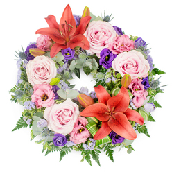 A meticulously arranged circular sympathy wreath fills the frame against a pure white backdrop, its composition both elegant and deeply comforting. Two prominent Asiatic lilies in a rich reddish-orange hue act as luminous focal points, one positioned near the top of the ring and the other closer to the lower curve. Their broad petals and delicate stamens create a sense of gentle radiance, like small suns within the design. Nestled around them are generous clusters of soft blush and lavender-pink roses, each bloom fully open and layered, giving the wreath a plush, velvety appearance. Interwoven between the roses are ruffled eustoma flowers, ranging from delicate pink to intense, regal purple, their cup-shaped blossoms adding depth and movement. Fine sprays of lavender-blue filler flowers, reminiscent of statice or limonium, weave through the arrangement like a light mist, softening transitions between colours. Small, steel-blue eryngium thistles introduce a subtle architectural edge, their spiky forms contrasting beautifully with the rounded petals. The base is built on a foundation of lush greenery, including silvery eucalyptus, feathery ferns, and varied glossy leaves that give the wreath a full, living presence. A few closed lily buds are nestled among the foliage, suggesting future blooms and the quiet continuity of love beyond loss. The overall design feels serene and composed, the kind of funeral wreath a family in Canonbury might place at a farewell service or deliver to a home near New River Walk, offering a dignified message of sympathy, remembrance, and support.