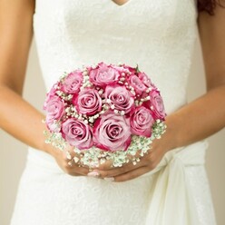 Resting serenely against the opulent lace of a Canonbury bride's gown, this bridal bouquet is a stunning study in floral artistry and gentle emotion. Each rose is a vision in itself, blending pale pink centres into vivacious magenta tips, their petals unfolding with a silken sheen that seems to glimmer with anticipation. Bathed in the soft daylight that streams through local Islington windows, the bouquet's rounded, compact form is enriched by wisps of snowy white baby's breath, lending a dreamy haze between the blooms. The bride's hands-warm and relaxed, nails reflecting a subtle, light shimmer-enfold the bouquet with clear devotion, mirroring the loving touch of the florist who arranged it. The gown, a sleeveless masterpiece adorned with intricate floral lace and a gentle V-neckline, brings tradition and modern grace together in harmony. Above her hands, a delicate sash or tie binds the composition. The blurred, neutral background intensifies the vividness of the flowers and the radiance of the moment. Crafted with care not far from Canonbury's elegant New River Walk, this scene epitomises the romance and singularity every couple hopes for-an unforgettable bouquet created for a once-in-a-lifetime occasion.