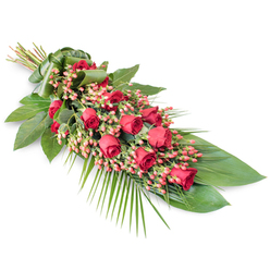A refined and dignified funeral spray stretches diagonally across a crisp white background, its elongated form drawing the eye from the neatly bound stems at the upper left to the tapered, softly fanned blooms at the lower right. This is a classic red rose tribute, the kind often chosen by Canonbury families for services near Upper Street or small, intimate memorials at local churches. Layers of velvety red roses line the centre, some still in bud with tight, promising swirls of petals, others partially or fully open, displaying lush, romantic faces that suggest deep affection and honour. Nestled among these roses are abundant clusters of small Hypericum berries, their round shapes tinted a gentle coral-pink to pinkish-red, like tiny lanterns adding delicate texture and subtle variation in tone. Beneath and around the flowers, a cushion of rich greenery provides structure: large, glossy leaves form a dark emerald base, shining gently as though polished, while slender, feathery palm fronds stretch out from the sides and lower edge, giving the whole design a sense of gentle movement and breath. Finer stems and wisps of foliage weave through the arrangement, filling any gaps and creating a layered, three-dimensional look. The composition feels thoughtfully balanced, neither heavy nor sparse, with enough negative space to appreciate each rose and berry. Against the white backdrop, every shade of red and green is amplified, transforming this sympathy spray into a visually striking yet serene farewell. It conveys a message of respect, remembrance, and comfort, suitable for delivery to a Canonbury funeral home, crematorium, or a quiet service held at a family home.
