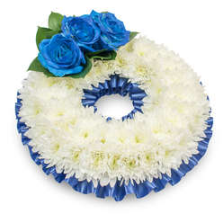 From a slightly elevated viewpoint on a pristine white field, this circular memorial wreath presents a composed, reverent silhouette. The main body is formed of a dense ring of white chrysanthemums; each bloom's delicate, feathery petals overlap to produce a lush, tactile surface with a subtle dome and soft cream touches at some centres that warm the white. A cluster of three deep electric-blue roses sits at the upper left, their velvety petals spiralled tightly and saturated with a calm, solemn blue that reads as both distinctive and soothing. These roses are cradled by glossy, dark green foliage which provides a natural counterpoint to the pale chrysanthemum ring. Both inner and outer rims of the wreath are finished with a pleated satin ribbon in matching cobalt blue - the ribbon's sheen and ruffles catch light and shadow, tracing the shape and lending a formal, thoughtful edge. The wreath rests with a faint shadow beneath it, suggesting presence and gentle elevation as if ready to be placed beside a casket or on a memorial easel. I picture this kind of tribute arriving from Canonbury Flowers for a service at a small family chapel near Newington Green or to be laid at a graveside in Barnsbury; its balance of white purity and dignified blue offers comfort and remembrance. The overall impression is one of serene reverence - a handcrafted emblem of sympathy intended to honour and console.