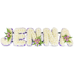A deeply personal memorial piece spells the name JENNA in large, three-dimensional block letters, each letter formed from tightly packed, creamy white chrysanthemums or similar small florets to create a soft, pillowy texture against a pristine white field. A slim, deep-purple trim-appearing as ribbon or tiny clustered blooms-outlines each letter's inner edge, sharpening the forms with an elegant, somber accent. Delicate clusters of flowers adorn key points of the design: a gentle spray on the upper curve of the 'J', a modest bouquet at the lower centre of the first 'N', and a third gathering atop the right side of the 'A'. Those accents are composed of blush-pink and lavender spray roses, with touches of light cream or pale yellow roses and feathery dark-green ferns that give a natural, living quality to the otherwise formal lettering. Thin dark metal or wooden bars subtly join the letters, providing a discreet structural spine suitable for display on an easel at a service. The overall impression is one of quiet dignity-soft petals, cool greens, and the calm contrast of purple trim that suggests remembrance and reverence. A faint, fresh floral scent and the tactile softness of the chrysanthemums help make this an intimate farewell. Prepared by local hands, this style of name tribute is well suited for funeral venues and wakes throughout Canonbury, gently conveying respect, memory, and the comfort of a carefully chosen floral goodbye. Canonbury Flowers can craft this same personalised tribute with local delivery across Canonbury and nearby Highbury and Essex Road.