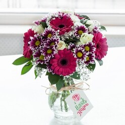 A richly textured bouquet of mixed flowers stands proudly in a squat, clear glass vase, centred against a luminous white backdrop that suggests a bright Canonbury morning streaming through a window. The arrangement is dominated by large gerbera daisies in intense shades of deep pink and magenta, their dark, almost chocolate centres drawing the eye straight into the heart of each bloom. Interwoven among them are generous clusters of chrysanthemum-like flowers, their petals a striking bicolour of deep violet at the tips that gradually softens to white, encircling radiant yellow centres that echo small bursts of sunshine. Creamy white roses, some gently unfurling and others wrapped in tight, elegant buds, add sophistication and a soft fragrance you can almost imagine. Wisps of delicate white baby's breath float between the larger blooms, lending a cloud-like lightness and enhancing the sense of airy fullness. At the base, rich green foliage and sturdy stems are visible through the water in the vase, their arrangement precise and deliberate, as you would expect from a skilled florist who regularly creates gifts for Canonbury townhouses and flats near Essex Road. The vase's neck is wrapped with simple rustic twine, tied into a neat bow that introduces a handcrafted, homely touch. Suspended from the twine is a small, rectangular white gift tag printed with a pink floral motif and subtle script, suggesting a thoughtfully written note for celebrations such as birthdays, congratulations, or a cheerful pick-me-up. The diffused light caresses each petal and leaf, making the colours appear freshly cut, as though the bouquet has just arrived via local flower delivery, ready to brighten a dining table or hallway console.