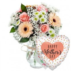 Set against a spotless white backdrop, this Mother's Day composition feels like a pause of calm joy amid the bustle of Canonbury's streets. A clear glass vase, gently tapered and finished with a simple wrap of rustic twine, holds a generously rounded bouquet that seems to glow from within. At the heart of the arrangement, peach-pink roses are opened to their full, velvety spirals, each petal curving softly as if cupped by hand. Alongside them, peach-orange gerbera daisies with dark, almost black centres bring a playful, daisy-like boldness, echoing the cheerful spirit of local weekend markets nearby. Threaded through the larger blooms are clusters of tiny white baby's breath, creating a misty halo, while small daisy-shaped chrysanthemums with light green centres add structure and a fresh, garden-picked feel. Glossy green leaves frame the flowers, giving the design a sense of depth and movement, as if a breeze has just passed through the bouquet. A slim white greeting card peeks shyly from behind the flowers, its colourful lettering mostly hidden, suggesting a private message of thanks and affection. Anchored beside the vase and leaning towards the arrangement is a large foil heart balloon. Its surface is bright white, scattered with sparkling gold and silver dots, and in the centre rests a soft peach heart emblazoned with the words "HAPPY Mother's DAY" in bold black lettering, the word "Mother's" flowing in elegant script. This carefully styled scene captures the emotion of a Mother's Day delivery arriving at a Canonbury home: warm appreciation, gentle celebration, and the promise of shared memories.