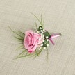 A single pink rose boutonniere is laid carefully on a softly textured, light beige linen cloth, the fabric's natural weave catching the gentle daylight like the table linens you might see at a Canonbury wedding reception. Viewed from a slightly elevated, eye-level angle, the rose is the clear focal point, its petals unfolding in a generous, full bloom. At the centre, the hue is a warm, blush pink that gradually deepens to a richer, more romantic magenta at the outer edges, each velvety petal curling delicately as if just opened that morning. Clustered closely around the base of the rose are sprigs of pure white baby's breath, their tiny, cloud-like blossoms forming a soft halo that gives the whole piece an airy, almost weightless feel. From behind the rose, fine, light green strands of decorative foliage - like delicate ferns or slender grasses - fan out in relaxed arcs, adding a sense of movement and a fresh, garden-picked character that feels right at home in the leafy streets near Canonbury Square and along St Paul's Road. At the base, the stems are carefully gathered and wrapped in a smooth pink satin ribbon, tied into a neat, understated bow that mirrors the rose's colour and lends a refined finish suited to a groom's or prom date's lapel. A slim silver pin is visible, tucked against the stems and ready to secure the boutonniere to a jacket. Soft, even lighting casts gentle shadows that reveal the three-dimensional structure of each flower and leaf, making this small arrangement feel quietly luxurious - the kind of thoughtful detail chosen for weddings, proms, or formal celebrations in and around Canonbury, where elegance is expressed in subtle, handcrafted touches.