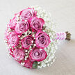 Resting at an elegant diagonal on a softly crumpled, light beige cloth, this bouquet feels like a quiet moment before a Canonbury wedding begins. A cluster of plush roses forms the centrepiece, each flower painted in a gradient of pinks and purples: the innermost petals are a muted lavender-pink, gradually deepening to vivid fuchsia and rich magenta at the edges. The blooms are in various stages of opening, from tight buds to gently unfurling spirals, creating a sense of life and movement within the rounded design. Surrounding them is a generous mist of white baby's breath, its countless tiny blossoms forming a frothy, ethereal halo that softens the strong rose tones and adds a light, airy texture to the arrangement. A few dark green leaves peek from beneath the flowers, offering a subtle, earthy counterpoint to the pastel and jewel-like hues. At the base, the stems are neatly aligned and bound with a smooth lilac satin ribbon, its surface slightly glossy, as though just tied by an experienced florist. A winding strand of small white pearls coils around the handle, mirroring the delicate gypsophila and giving the bouquet a classic, ceremonial finish. The neutral background fabric, reminiscent of fine table linen in a local Canonbury restaurant or a reception venue near Upper Street, keeps the eye entirely on the flowers. This composition captures the romance and poise ideal for a bride, bridesmaid, or special anniversary celebration, and suits intimate local gatherings as well as elegant events across Islington.