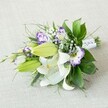 A hand-tied bouquet of white lilies and purple lisianthus rests diagonally across a softly textured, light beige fabric, as if placed on a linen runner in a Canonbury townhouse. The composition is long and slightly tapered, with the lush flower heads fanning out on the left and the bound stems pointing to the right. At the heart of the design, several large Oriental lilies are fully open, their creamy white petals curving back to reveal golden-yellow stamens dusted with pollen and a fine constellation of reddish-brown speckles. Around them sit plump chartreuse-green lily buds, closed but full of promise, their smooth surfaces catching the gentle, diffused light. Threaded between the lilies are ruffled lisianthus blooms, their bell-shaped petals edged in vivid purple that melts into clean white throats, some in deeper violet tones, others a soft lavender haze. Wisps of airy white gypsophila float through the design like tiny clouds, weaving a delicate veil between the larger blooms. A rich variety of greenery frames the flowers: broad, glossy dark green leaves give weight and structure, while finer, spiky and feathery fronds add movement, as if the bouquet had just been lifted from New River Walk. The stems are neatly wrapped in a crisp white satin ribbon, embellished with tiny pearl-like beads that catch the light along the narrower end, hinting at a bridal bouquet made ready for a Canonbury ceremony. The overall mood is one of fresh elegance and quiet celebration, perfectly suited to a wedding, anniversary, or meaningful gift delivered from a trusted local florist in N1.