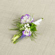 A compact celebration of spring, this boutonniere combines cool white lisianthus edged in soft violet with airy accents that breathe life into the composition. Two lisianthus heads sit as the focal anchors, their ruffled, satin-like petals catching the light and offering a subtle perfume; around them, miniature sprays of baby's breath float like tiny constellations, providing a delicate halo of white. Narrow flutes of grape hyacinth introduce a lively blue-violet intensity-short, clustered spikes that contrast the lisianthus' rounded bloom and add an almost musical rhythm to the silhouette. Featherlike fern tendrils slip through the arrangement, lending a woodland freshness and an elegant asymmetry that reads beautifully on a lapel. The stems are finished with a neat white ribbon, chosen to echo the petal tones and to sit unobtrusively beneath a jacket. Set against textured linen under soft daylight, every surface-petal, leaf, ribbon-reveals gentle highlights and the tactile pleasure of silk-fine petals. As a local florist who often supplies grooms and guests for ceremonies around Essex Road and the New River Walk, I picture this pinned for a Canonbury wedding or an intimate celebration on a side street near De Beauvoir; it's small but purposeful, designed to be noticed in close-up photographs and to complement refined attire. Prepared with the attention to proportion and scent that customers from Canonbury expect, this boutonniere is offered by Canonbury Flowers as an elegant finishing touch for the day's most treasured moments.