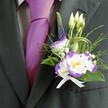 A close, carefully framed photograph shows the upper torso of a man dressed for a smart Canonbury wedding, the focus resting on his refined buttonhole and coordinated formalwear. He wears a dark charcoal grey suit jacket with a clean, tailored line, the fabric catching soft, indoor light so that the texture of the weave feels almost touchable. Beneath, a light grey checkered shirt introduces a gentle pattern, while a lustrous magenta silk tie sits perfectly knotted at the collar, its rich colour echoing the purple tones in the flowers. Pinned neatly to the left lapel is a thoughtfully crafted boutonniere: a main lisianthus bloom of crisp white petals edged in deep violet, accompanied by a second partially open flower to one side. Around them cluster small, closed white buds, tiny sprigs of baby's breath, and a slender spire of dark indigo-purple veronica that lifts the eye upward. Wisps of feathery greenery and broader emerald leaves create layers of texture, with looping strands of fine decorative grass adding a modern, almost architectural curve. At the base, a small white satin ribbon is tied into a precise bow, its sheen mirroring the shine of the tie. The composition feels elegant yet relaxed, the kind of buttonhole a groom might wear for a ceremony near Canonbury's leafy New River Walk or a celebration in one of its intimate townhouses, capturing the atmosphere of fresh beginnings, careful craftsmanship, and quiet joy that lingers throughout the day.
