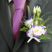 Resting just above the heart on a deep charcoal lapel, this boutonnière offers a composed blend of classic romance and contemporary detail that would suit any elegant occasion in Canonbury or nearby Islington. The focal white lisianthus, edged in striking purple, opens like a small, luminous moon; beside it a compact dianthus - white with lively purple ruffles - adds playful texture and floral punctuation. Wisps of baby's breath (gypsophila) scatter around the central blooms, creating a soft halo that suggests morning mist over the New River Walk. Tall, narrow veronica in deep indigo provides a vertical accent that lifts the eye, while feathery ornamental grasses and rich green foliage shape the silhouette and lend movement. A trio of slender lisianthus buds rises behind the main bloom, hinting at continuity and new beginnings; the arrangement is tied with a crisp white ribbon whose clean fold complements the shine of a worn magenta tie. Light skims the petals, sharpening the purple edges and revealing velvety textures; the gentle, green-sweet scent of fresh florals offers a comforting undercurrent throughout a long celebration. As crafted by a local Canonbury florist, the composition feels personal and considered - perfect for a groom, best man, or honoured guest who wants an accessory that reads both tender and assured. It's the kind of small living artwork that people notice in photos and remember in conversation long after they leave the reception near Essex Road or stroll past St. Paul's Road.