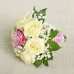 A carefully arranged petite bouquet lies diagonally across a softly textured, light beige fabric that resembles finely woven linen, the neutral tone allowing every flower to stand out clearly. At the centre, five rounded ivory roses unfurl in full, layered petals, their creamy colour evoking the gentle calm of a quiet Canonbury morning. Nestled near the front-left of the grouping is a single rose in a vivid shade of fuchsia pink, its saturated tone drawing the eye and adding a playful yet elegant burst of colour among the classic whites. Interlaced throughout the roses are frothy sprays of white baby's breath, each tiny blossom forming part of a delicate, cloudlike surround that adds height and airiness without overwhelming the design. Around the edges, deep green pointed leaves, likely ruscus or salal, extend just beyond the blooms, giving contrast and depth, much like the leafy borders along the paths near Canonbury Station. The stems are gathered tightly and wrapped in a glossy bright pink satin ribbon, forming a short, comfortable handle. Over this ribbon, multiple strands of small, lustrous white pearls are arranged in precise horizontal rows, their gentle sheen lending a refined, almost heirloom quality that suggests the care of an experienced florist. The lighting is soft and even, without harsh shadows, emphasizing the natural textures of petals, leaves, and fabric. The overall composition feels intimate and romantic, perfectly sized for a wrist corsage, boutonniere, or a small hand-tied posy-ideal for weddings, school leavers' events, or special dinners in the Canonbury area where subtle elegance is cherished.