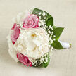 On a pale, slightly crumpled linen backdrop the colour of warm oatmeal, a romantic bouquet lies horizontally as though it has just been set down between photographs at a Canonbury townhouse wedding. The central bloom, a generous creamy white peony, is open and luxurious, its many finely ruffled petals forming a soft, cloud-like sphere that draws the eye immediately. Clustered around this focal flower are vivid fuchsia-pink roses, each bud tightly layered and velvety, adding depth and a classic, romantic tone to the arrangement. Wandering delicately between the larger blooms are wisps of white gypsophila, or baby's breath, their tiny blossoms creating a lacy halo that softens the transition between the bold colours and the neutral fabric underneath. Encircling everything, sweeping arcs of deep green leaves form a graceful frame, their glossy surfaces catching the gentle light in much the same way leaves do along the shaded paths of Highbury Fields on a mild day. The stems of the bouquet are carefully gathered, visible at one end and wrapped with a neat band of white satin ribbon that suggests a florist's practiced hand and hints at a bride's steady grip. The lighting in the image is calm and diffused, without harsh shadows, allowing the creamy whites, rosy pinks, and rich greens to appear natural and fresh, as if prepared that morning at a Canonbury florist studio. The mood is one of poised anticipation, suitable for a wedding, civil ceremony, or romantic celebration, where the bouquet's refined blend of texture, colour, and softness can be fully appreciated.