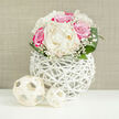 A romantic, dome-shaped floral arrangement of ivory peonies and vivid fuchsia-pink roses is showcased in a modern white basket-style vase, as if waiting on the sideboard of a calm Canonbury flat just off Essex Road. The peonies are lush and full, their creamy petals unfolding like layers of silk, hinting at a soft, powdery fragrance that suggests bridal mornings and freshly pressed linen. Nestled alongside them, the pink roses offer a deeper, more saturated hue, each layered bloom suggesting affection, gratitude, and a touch of drama. Threaded gently through the larger flowers, clouds of white baby's breath add a gauzy, almost mist-like texture, softening the bold forms and giving the bouquet a dreamy, romantic glow under the bright, even lighting. Around the base, smooth, broad green leaves arc outward, framing the florals and grounding the palette with a rich, natural green. The flowers are gathered into a distinctive round white vase crafted from thin, interwoven strands that resemble painted rattan or slender branches, its open lattice design allowing glimpses of stems and shadows inside. On the sleek white surface in front of the vase lie two small woven spheres in a pale wood tone, their airy construction echoing the vase and adding a touch of minimalist design. The subtle beige backdrop, like a softly textured fabric wall, recedes quietly so the blooms and textures can shine. The overall effect is of gentle sophistication and quiet celebration, perfect for a registry reception near Islington Town Hall, an anniversary dinner at home, or same-day wedding flower delivery across Canonbury and neighbouring De Beauvoir.