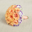 Seen from a slightly lifted viewpoint, this elegantly rounded bouquet rests across a soft, textured beige fabric that emphasizes the warmth of its flowers. The heart of the arrangement is a cluster of abundant roses in peach and soft orange, their petals layered like small suns opening toward the light. Among them, crisp white lisianthus with violet-edged ruffles introduce a cool counterpoint, their distinctive colouring tracing each petal's silhouette and lending the bouquet an artful depth. Fine sprays of baby's breath float through the design, creating gentle pockets of air that make the heavier roses appear to breathe. The stems are wrapped in a silk-like satin ribbon in a muted coral-peach hue, and a delicate strand of cream pearls curls around the handle in a serpentine line, a refined touch that hints at vintage jewelry and quiet luxury. The overall texture moves from velvety rose faces to feathery gypsophila and ruffled lisianthus, giving the composition both visual and tactile variety. Bathed in soft daylight, the bouquet's colours read warm and inviting-ideal for a Canonbury wedding beneath plane trees or a small celebration on a summer evening near Essex Road. This design feels intimate and joyful, balanced between traditional romance and a contemporary twist, the kind of bouquet chosen by couples who want warmth, personality, and thoughtful craftsmanship in every bloom.