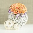 A sophisticated floral display rests on a pristine white surface, evoking the calm charm of Canonbury's tree-lined squares and period homes. The arrangement centres on a luxuriant dome of roses in gentle peach and apricot tones, each bloom full and softly rounded, with petals layered in a way that suggests both warmth and careful craftsmanship. The roses are packed closely into a near-perfect sphere, giving a sense of generous abundance while maintaining a crisp, sculpted outline. Nestled at the foot of this rose globe is a surrounding band of white lisianthus, their ruffled petals traced with a vivid band of purple, creating a striking yet elegant contrast that feels modern but not overpowering. Little clouds of white baby's breath flicker between the lisianthus blooms, lending lightness and a slightly whimsical touch, like tiny stars sparkling at the edge of dusk. The flowers rise from a rounded white woven vessel, its broad, bulbous form fashioned from overlapping wicker or rattan strips that weave together into an open, airy lattice. This handcrafted container echoes the artisanal details you might find in a carefully curated Canonbury interior, while its bright white finish keeps the focus firmly on the flowers themselves. In front of the centerpiece lie two decorative wooden spheres in natural beige tones, one larger, one smaller, their fine woven strands creating delicate shadows on the surface below. A softly textured beige fabric backdrop completes the setting, holding everything in a warm, neutral embrace. The effect is romantic, polished, and quietly joyful, ideal for wedding table arrangements, anniversary dinners, or serene home styling in North London.