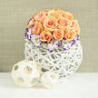 Bathed in soft daylight, this rounded bouquet forms a compact, dome-like mass of roughly two to three dozen apricot and soft-peach roses whose layered petals create a lush, tactile surface. You can sense both heady fragrance and the gentle give of petals as fully opened blooms sit alongside tighter buds, producing depth and movement across the composition. Nestled around the roses are white Lisianthus flowers, their creamy faces edged with a lively indigo to deep-purple rim that supplies a refined, painterly accent against the warm rose tones. Throughout the bouquet tiny wisps of Gypsophila (baby's breath) float like lace, introducing an ethereal softness that balances the roses' rich fullness. The stems are arranged in a contemporary white woven orb vase-an airy lattice of reed or wicker painted bright white-its openwork silhouette suggesting handmade craft and letting discreet shadows fall onto the tabletop. To the left, two decorative wooden spheres constructed from thin bamboo strips repeat the woven motif and add a tactile, sculptural counterpoint. Behind the arrangement, a fine-textured, natural linen-coloured wall is gently out of focus, making the floral colours appear more vivid and intimate. I imagine this piece on a mantel or dining table in Canonbury, placed for a small wedding or celebratory dinner near New River Walk or Essex Road market; it offers the composed romance and understated luxury of thoughtful floral design, the kind a local florist would create for anniversaries, housewarmings, or simply to lift a morning room with quiet, seasonal cheer. Canonbury Flowers often uses similar palettes when styling elegant, neighbourhood events.