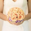 A close-up wedding photograph shows the midsection of a bride standing in soft, natural light, her hands gently cupping a rounded bridal bouquet that seems to glow against the creamy white of her gown. The dress is classic and romantic, with delicate lace tracing the bodice and a modest V-neckline, the fabric falling in soft, graceful folds that blur into the background. At the centre of the image is a lush cluster of roses in tender shades of peach, apricot, and blush pink, each bloom tightly petalled and velvety, forming a perfectly domed shape that feels both abundant and carefully controlled. Just beneath this cloud of warm colour, a ring of deep purple flowers edged in white-likely lisianthus-peeks out, their ruffled petals adding drama and contrast, like a shadow framing the light. Tiny sprays of white filler flowers, reminiscent of baby's breath, weave around the base and between the blooms, softening the edges with a mist-like texture. The bride's nails are neatly manicured in a subtle nude or pale pink tone, one thumb catching a glint of shimmer that echoes the quiet luxury of the bouquet. The overall mood is serene and anticipatory, capturing the stillness of a Canonbury bride moments before walking through a leafy garden or into a townhouse ceremony. You can almost imagine the gentle fragrance of fresh roses lingering in the air, the bouquet created by a local florist who understands how to balance romance, colour harmony, and personal style for weddings along the quiet streets and squares of this North London neighbourhood.