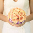 This arrangement reads like a soft poem for a wedding morning: the roses, gathered in a symmetrical, almost architectural cluster, glow in apricot warmth, each bloom unfurling in smooth, silken spirals that catch the gentle studio light. Around this sunlit centre, a ring of purple-tipped lisianthus offers an elegant, cool contrast - like twilight clouds hovering around a peach horizon - their slender stems and ruffled edges giving the design a refined, botanical lace. Interspersed among the main blooms, a delicate lattice of white baby's breath and fine green sprigs adds an airy filigree that lightens the bouquet's silhouette and suggests a weightlessness suited to a bride's embrace. The photograph emphasises texture: the creamy density of rose petals against the translucent edges of the lisianthus, all softened by diffused light that renders shadows soft and petals almost luminous. The scent is imaginable here - a fresh, sweet rose perfume with faint green notes - conjuring a calm, expectant morning before vows. This bouquet feels both contemporary and classically bridal, composed with precise attention to shape and rhythm so it sits naturally against lace or silk. For Canonbury couples exchanging vows near Newington Green or on Essex Road, the palette and scale read as quietly celebratory and deeply personal, intended to be the gentle focal point of ceremony photographs and a keepsake of the day's promises.