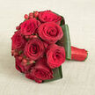 On a warm, linen-like beige surface lies a rounded bouquet of deep red roses, designed with the kind of meticulous care you might expect from a Canonbury florist preparing flowers for an intimate town hall wedding. The roses form a compact, almost sculptural dome, each head snugly nestled against the next, so their layered petals create sweeping whorls of crimson and dark burgundy. Some blooms are more open, revealing their intricate centres, while others remain slightly closed, promising more beauty to unfold. Punctuating the sea of red, small sprays of glossy hypericum berries peek through, their brighter scarlet hues and smooth, bead-like shapes adding texture and a subtle sparkle. Encircling the entire posy is a collar of deep green leaves, broad and neatly folded, their edges aligned with a dressmaker's precision to give the bouquet clear contours and a contemporary finish. The handle, extending to the right, is wrapped in vivid red satin ribbon, secured so seamlessly that it creates a sleek, comfortable grip for a bride or partner holding it along the tree-lined streets around Canonbury Square. The light in the image is soft and even, with no harsh shadows, allowing viewers to appreciate every velvety petal and reflective berry. It feels timelessly romantic, like a bouquet chosen for a winter wedding, a golden-hour photo shoot by the Regency terraces, or a heartfelt anniversary surprise delivered across Canonbury, Highbury, and nearby neighbourhoods. The overall impression is one of opulent, focused passion, refined by expert craftsmanship.