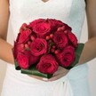 In the gentle ambience of a Canonbury morning, a bride's delicate hands cradle a classic wedding bouquet that radiates romance and warmth. The central feature is an opulent, rounded cluster of deep crimson roses, their petals velvety and rich, some tightly coiled in bud, others softly blooming, each a promise of love's continuity. Dotted among the roses, polished clusters of scarlet hypericum berries emerge, serving as playful, jewel-like interruptions that add a festive, tactile dimension to the visual poetry. Encasing the vibrant reds, a collar of broad, glossy green leaves gives the bouquet defined structure and botanical sophistication, echoing the lush gardens of Canonbury Square. The bride's white gown, adorned with fine lace patterns and a gentle drape visible at the skirt, forms a luminous canvas that makes the red hues almost luminous. Soft, natural light washes over the scene, drawing out the bouquet's textures and lending a radiant, celebratory feel - as if the moment could linger forever along the leafy lanes near Canonbury station. This bouquet, crafted with expert care by local florists, encapsulates the joy, excitement, and tradition that define wedding days in Canonbury, creating a lasting symbol of love and local artistry.