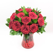 A classic dozen red roses stands proudly at the centre of this image, the blooms arranged in a tight, rounded dome that fills the frame against a flawless white studio background. Each rose is a deep, velvety crimson, the petals layered in perfect spirals that curl inward to form rich, romantic hearts. The texture appears soft and plush, as if each flower has been freshly opened that morning, with no bruising or blemish to disturb the smooth curves. Between the roses, glossy dark green leaves, likely Ruscus, peek through, their pointed shapes and reflective surfaces adding freshness and contrast to the intense red. The foliage gathers more thickly around the base of the bouquet, cradling the flowers and giving the arrangement a finished, polished edge. All of this is set in a translucent ruby-red glass vase with a gentle, fluted silhouette, slightly wider at the top and base and slimmer through the middle. The glass shows subtle swirling or ribbed detailing that catches the studio lights, creating faint highlights and shadows that echo the natural texture of the petals. Through the tinted glass, the straight, dark stems are visible, crossing and overlapping in organic lines that anchor the design. The bright, even lighting gives the whole composition a clean, contemporary feel while still allowing the arrangement to exude warmth, intimacy, and passion. It looks like the kind of romantic bouquet a Canonbury resident might send across N1 for a Valentine's surprise, an anniversary dinner near Highbury Corner, or a heartfelt declaration delivered by a local florist who understands the language of classic red roses.