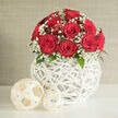 A lush cluster of red roses is gathered into a near-perfect dome, each flower at its fullest, with velvety petals that curl inward in tight, elegant swirls. Around the roses, a halo of pure white baby's breath wraps the bouquet in a soft, mist-like border, small blossoms forming frothy clusters that lighten the overall look and accentuate the richness of the red. A few glossy green leaves emerge discreetly between flower heads, adding depth and a touch of natural contrast without disrupting the rounded silhouette. The arrangement is seated in a distinctive spherical vase, woven from slender white strips that crisscross to create an airy, open structure reminiscent of painted rattan or wicker. Light filters through the gaps in this sculptural vessel, creating subtle patterns on the clean white tabletop below. Next to the vase lie two decorative woven balls in complementary cream tones, one medium and one small, their textured surfaces and simple shapes mirroring the vase's craftsmanship and adding to the curated, modern feel. The background is a softly blurred wall in neutral beige-grey, quietly supporting the composition so the focus remains firmly on the interplay between passionate red blooms and calm white surroundings. The lighting is soft and even, as if caught on an overcast afternoon in a Canonbury apartment overlooking New River Walk, perfect for highlighting an arrangement chosen as a wedding centrepiece or romantic gift. It feels both contemporary and timeless, like something a local florist would design for a chic registry office ceremony or an intimate reception at a nearby Islington venue.