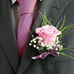 Pinned neatly to a charcoal-grey lapel, this pink rose boutonniere feels like a quiet love note worn close to the heart, the kind of detail you might notice at an intimate Canonbury ceremony by the New River Walk. The rose itself is a soft, luminous pink, opening in smooth, layered petals that curl gently at the edges, as if in a slow, graceful breath. Around it, delicate sprays of white gypsophila-baby's breath-form light, cloud-like clusters, their tiny blossoms adding a sense of innocence and airy romance. Finely textured greenery, with slender, feathery leaves, fans out behind and alongside the rose, introducing depth and freshness, as though a piece of a well-kept garden has been gathered and refined for this single moment. The stems are bound with a lilac satin ribbon, tied with practiced precision; the ribbon's sheen adds a refined, celebratory touch that harmonises beautifully with the formal suit fabric. Subtle, natural light skims across the petals and lapel, accentuating every curve and thread, making the rose appear almost dewy and newly picked, as if just prepared by Canonbury Flowers for same-day wedding delivery. You can imagine a faint, floral fragrance rising each time the wearer moves, blending with the crisp scent of greenery. This boutonniere is more than an accessory-it is a crafted expression of respect, warmth, and commitment, perfect for weddings, civil ceremonies, or special occasions where style and sentiment are meant to be remembered long after the day has ended.