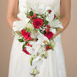 Held gently at waist height against a white wedding gown adorned with subtle lace and embroidered details, this cascading bridal bouquet unfurls like a river of red and white blossoms flowing through a quiet Canonbury side street on a wedding morning. At its heart are generous crimson roses, their layered petals plush and velvety, conveying a depth of romance that feels both timeless and deeply personal. Around them, elegant white lilies open wide, their crisp petals and visible green stamens adding freshness and a sense of serene composure, as if they have just been gathered from a nearby Islington flower stall. The bouquet lengthens into a graceful waterfall, where white Phalaenopsis orchids trail downwards in soft arcs, their butterfly-like blooms and delicate buds shining faintly in the gentle light. Interlaced among these statement flowers, sprays of tiny white baby's breath create a light, airy veil, softening the reds and echoing the bridal tulle. Small, gleaming red Hypericum berries nestle within the foliage, bringing playful dots of colour and texture, like scattered confetti along the New River Walk. Lush greenery in varied shapes and tones-slender fronds, rounded leaves, and subtle green-tipped white florets-frames the bouquet and gives it structure. The neutral, softly blurred background in warm cream keeps all attention on the flowing teardrop design, making the image a sensory-rich portrait of a classic red-and-white wedding bouquet, ready to be carried down the aisle of a local Canonbury church or registry office.