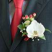 A refined close-up of formal wear, expertly balanced between fashion and floristry: a pristine white Phalaenopsis orchid is pinned to the left lapel of a dark charcoal suit, its broad petals smooth and almost silky to the eye, the central labellum marked with delicate yellow-gold and white patterning that gives the bloom a quietly radiant centre. Surrounding the orchid, glossy deep-green foliage provides weight and structure, while sprays of gypsophila float like a gentle mist, lending ethereal softness and a whisper of old-fashioned romance. Nearby, plump red hypericum berries offer a warm, coral-tinged contrast that mirrors the vibrant red satin tie knotted sharply against a light patterned dress shirt - the tiny grid or check visible at the collar. A slender twist of matching red ribbon secures the stems, echoing the tie's sheen and tying the look together with polished restraint. The charcoal fabric of the suit shows a fine tailored texture under soft, natural light, creating subtle shadows that highlight the boutonnière's three-dimensionality. As a local florist in Canonbury, Canonbury Flowers would arrange this for a bridegroom, prom guest, or gala attendee wanting a clean, modern accent with classic sentiment. The composition invites touch: the orchid's cool velvet, the berries' firm roundness, the ribbon's satiny slide - sensations that pair with emotions of anticipation and quiet joy on a day centred around family, friends and ritual in the nearby streets of Essex Road and New River Walk.