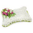A long, rectangular pillow of blooms sits serenely against a clean white backdrop, its surface completely covered in tiny, snow-white chrysanthemums that read like a plush textile. The dense, button-like faces of the chrysanthemum create a soft, clouded texture you can almost feel beneath your fingertips, suggesting purity and gentle repose. At the top-left corner a concentrated spray of deep magenta and fuchsia roses draws the eye - their velvety petals opening in layered spirals that contrast richly with the pristine white field. Tucked among the roses are small clusters of pure white freesias or similar delicate buds, their slender stems and bell-shaped blooms offering a lighter, fragrant counterpoint. Lush green foliage, including narrow fern fronds and glossy leaves, fans out beneath the accent cluster and frames it against the pillow, adding cool depth and a living freshness. Around the pillow's edge a pleated loop of pale green satin ribbon peeks out like a soft border, catching the light and giving subtle definition to the rectangular shape while reinforcing the arrangement's garden-fresh palette. Soft, even studio light washes the piece, leaving a gentle shadow beneath that emphasizes its three-dimensional form and invites quiet contemplation. This traditional funeral pillow is presented with careful craftsmanship - a visual lullaby meant to offer comfort, dignity, and remembrance at a graveside or memorial service. For those in Canonbury seeking a composed and respectful tribute, the arrangement conveys peaceful sleep and enduring affection through texture, colour, and scent, speaking tenderly where words may fall short.