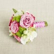 A small, hand-tied bouquet lies horizontally on a softly textured light beige fabric, the weave of the cloth recalling a simple linen table set in a Canonbury townhouse near Highbury Fields. At the centre of the arrangement, two fully opened pink roses and a third just behind them form the romantic focal point, their petals moving from a gentle blush at the heart to richer magenta tones tracing the outer edges, like watercolour shading. Nestled closely around them are neat pom-pom clusters of vivid lime green button chrysanthemums, their tiny packed petals creating a playful contrast in both texture and colour. Between the roses and mums, airy stems of creamy white filler flowers, likely lisianthus or delicate freesia, soften the structure, with a few tiny star-shaped white blooms with yellow centres adding an almost bridal sparkle. Hints of fresh green foliage peek through, anchoring the composition in nature. The stems are trimmed short and bound into a compact posy, wrapped in a smooth fuchsia pink satin ribbon that gleams gently in the soft, even light. Over the ribbon, a strand of small white faux pearls is wound diagonally, crossing and looping to create the impression of jewellery on the handle, while the bare stem tips show just beyond. The gentle lighting casts almost no harsh shadow, allowing every ruffle of petal and thread of fabric to remain visible. The overall impression is of a romantic, modern bridal or bridesmaid bouquet, the sort that might be carried into a Canonbury ceremony at a local church or intimate registry office celebration, radiating freshness, charm, and understated elegance.