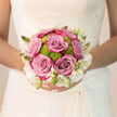 Held close against the bride's gown, this Canonbury bridal bouquet has the poise of something carefully composed, yet it still feels as natural as a stroll through nearby Highbury Fields at first light. At its heart, six classic roses in a soft shade between lilac and dusty pink unfurl in flawless spirals, each bloom plush and resilient, inviting the fingertips to trace their finely layered petals. Tucked between these roses sit cheerful lime-green button chrysanthemums, their rounded, textured heads bringing a lively modern twist and an almost playful energy to the arrangement. Encircling everything, a ring of petite creamy-white flowers and slender buds forms a subtle, lacy halo, reflecting the delicate detailing you might find on a wedding dress chosen from a boutique just off Essex Road. The colours move harmoniously from muted mauves to bright greens and gentle ivory, creating a bouquet that looks both contemporary and timeless, ideal for a Canonbury or Islington wedding. Soft daylight seems to skim across the blossoms, gently highlighting the curves of each petal and the crisp edges of the foliage beneath. In your mind's nose, you can sense a blend of fresh rose perfume and green, slightly herbal notes from the chrysanthemums and leaves, a scent that soothes while it uplifts. The stems are hand-tied and neatly bound, shaped to fit the bride's hands comfortably without overwhelming her silhouette. This is the kind of thoughtful design an experienced local florist would craft to stay beautiful from the moment she steps out onto a quiet Canonbury street to the final photographs under twinkling evening lights.