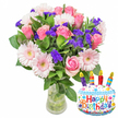Set against a clean white studio background, this celebratory bouquet reads as a quiet, joyful party in bloom. At its heart are abundant soft pink roses, their velvety petals unfurling from blush to a deeper pink, arranged so each bloom catches the light and suggests tenderness and warmth. Interspersed among the roses are pale pink Gerbera daisies with broad, smiling faces and buttery yellow centers that lend a playful, open energy. Small clusters of vivid purple filler-statice or limonium-are woven through the stems, their papery, clustered texture providing lively contrast and depth. A few tiny white spray blooms peek through, brightening the composition, while glossy deep-green leaves cradle every blossom and add a fresh, structured frame. The flowers sit in a clear glass vase with a gently widened base; the green stems and water are visible, a subtle reminder of freshness and careful florist technique. To the lower right, a metallic cake-shaped Mylar balloon punctuates the scene: a white 'frosted' top edged with wavy blue icing, scattered multi-coloured sprinkles, and four striped candles-purple, red, orange, blue-tipped with yellow flame motifs. Happy Birthday is printed across the front in cheerful, multicoloured letters, and a small ''KALEIDOSCOPE'' logo appears at the balloon's base. The overall effect is affectionate and festive-an arrangement that smells faintly sweet, catches the morning light, and balances romance and celebration-perfect for sending warm birthday wishes across Canonbury, Angel, or nearby Islington with thoughtful florist care.