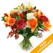 A hand-tied autumn bouquet arranged upright against a clean white background, composed with the quiet confidence of a neighbourhood florist. Large, trumpet-shaped orange lilies take centre stage, their wide-open petals and visible stamens casting a warm, sculptural presence; a few still-closed lily buds promise the bouquet will deepen over the coming days. Surrounding them are plump Gerbera daisies in ruby red and bright orange, their concentric petals forming cheerful, sunlike faces. Soft clusters of creamy white Lisianthus with gently ruffled edges introduce a cool, silky contrast to the bolder tones, while delicate Astrantia with purple-pink starry centres sprout fine, lacy texture between the larger blooms. Beneath the flowers, a bed of deep green foliage - ruscus or salal - gives the composition weight and a glossy backdrop, and strategically placed, deeply lobed oak leaves in burnt orange and reddish-brown frame the arrangement with an unmistakable autumnal silhouette. The stems are neatly gathered and bound with dark twine or raffia, the tactile knot visible at the bouquet's base and suggesting a freshly cut hand-tied style. In the lower right corner of the image a bright orange diagonal banner overlays the photograph, clearly reading FREE CHOCOLATE in bold white letters - a visible promotional detail rather than part of the flowers themselves. The overall impression is of a bouquet that smells faintly of green stems and autumn spice, ideal for sending warm thanks, a birthday surprise or a seasonal pick-me-up. Locals in Canonbury will recognise the easy, crafted style often chosen for deliveries around Canonbury Square and nearby streets, a composition made to sit warmly on a mantel or to greet a friend at their door.