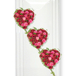 Three sumptuous hearts made entirely from roses descend gracefully down a spotless white wall, forming a striking and romantic funeral tribute that feels both contemporary and timeless. Each heart is cradled in dark ivy and built from an astonishing density of blooms; the roses are pressed close together so that the viewer sees a continuous sea of petals in shades of plush crimson, vivid magenta, and soft blush pink. The richer red tones sit at the core of each heart, speaking of passion, loyalty, and deep attachment, while the lighter pink roses brighten the design and bring in tenderness and gentle farewell. The ivy, with its slender stems and small, glossy leaves, outlines each heart like a frame and then trails downward to link one to the next, suggesting a continuous line of love that carries through generations. Set against a clean white backdrop that recalls the calm interior walls of a Canonbury chapel or a peaceful Islington ceremony space, the colours appear especially vivid, as though freshly misted and carefully lit. The hearts tilt ever so slightly on the diagonal, creating movement and a soft, cascading rhythm rather than a rigid column. This image evokes the feeling of a highly considered tribute crafted by an experienced local florist for a funeral in Canonbury or nearby Highbury Fields, perhaps placed near the front of a service where family and friends can gather close. The overall impression is of profound affection and remembrance, expressed through the enduring beauty of classic red and pink roses woven together with living greenery.