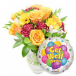 A radiant Get Well Soon ensemble set against a clean white backdrop, presented as if described by a local Canonbury florist standing in morning light. On the left a full, glass vase cradles a lush bouquet: large, opening roses in warm orange and deep yellow form the heart of the composition, their velvety petals catching a soft citrus-tinged scent and unfurling in layered spirals. Bright yellow gerbera faces lift like small suns, their textured centres adding a cheerful punctuation, while deep burgundy-to-maroon carnations provide ruffled contrast and sculptural depth. Sprays of delicate white alstroemeria thread through the arrangement, adding a breathy, porcelain-like lace that softens the stronger colours. Glossy green foliage with varied leaf shapes frames each bloom, giving the bouquet a garden-fresh fullness and a cool, leafy perfume. Stems and water are visible through a clear glass vase, the light refracting and showing the careful placement of each stem. To the lower right, slightly overlapping the floral mass, a round silver Mylar foil balloon glints with playful reflection. Its centre shows a cluster of cartoon-style balloons in bright blue, purple, green, pink and yellow, patterned with stripes, dots and checks, and bold yellow-and-blue lettering cheerfully reads Get Well Soon. Small starbursts embellish the white border and a tiny KALEIDOSCOPE logo sits along the bottom rim. The overall impression is one of warm encouragement and careful craft - a comforting, uplifting gift that would travel well from Canonbury to nearby Upper Street and Islington Green to brighten someone's recovery with colour, scent and sincere intent.