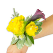 A vibrant wrist corsage rests gently on a light-skinned forearm against a crisp white backdrop, every element arranged to read clearly from the camera. The focal pair are two buttery yellow roses - the left bloom opened wide, the right a slightly smaller, cupped bud - each given a tiny clear rhinestone at its heart that catches the light like a secret glint. Set just behind and above the roses a single, slender calla lily in a deep plum-to-black-cherry tone unfurls its smooth, trumpet-shaped petal, introducing a dramatic counterpoint to the sunny roses. Surrounding foliage is generous but airy: fine, feathery asparagus fern lends movement and a soft, green halo; broader variegated leaves with pale, almost creamy edges (reminiscent of pittosporum) form a structured base; and clusters of miniature white filler flowers - small spray mums or tiny baby's breath - tuck into the shadows to add fullness. The corsage is mounted diagonally across the wrist from the photo's bottom-left to the middle-right, the fastening band mostly concealed so the flowers appear to float. Light is clean and slightly cool, emphasising petal texture and the glossy sheen of leaves; one can almost imagine the faint, warm scent of fresh roses mingling with the clean green notes of fern. This wearable arrangement reads as both celebratory and refined, equally suited to a prom, bridesmaid, or a guest arriving for a Canonbury Square ceremony, and reflects the careful hand of a local floral stylist attuned to colour, balance and the practicalities of on-the-wrist construction.