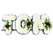 The image presents a bespoke sympathy tribute spelling TOM in large block capitals, the sort of arrangement often delivered to funerals and memorials around Canonbury. Each sturdy letter is completely surfaced with tightly packed white chrysanthemums, their many petals overlapping to form a thick, velvety blanket of white, giving the name a soft but clearly defined shape. Along the outer contour of every letter runs a royal blue satin ribbon, pleated and pinned in a precise line so that the rich blue colour frames the white flowers and adds depth, much like a calm strip of sky at dusk. At three main points-the upper left arm of the 'T', the outer curve of the 'O', and the top right of the 'M'-small floral sprays break the white expanse. In each spray, a few deep crimson red roses open fully, their petals velvety and layered, surrounded by smaller white or cream spray roses that soften the contrast. Delicate white, freesia-like blooms arc gracefully among the roses, while fresh green fern and glossy foliage spill around the edges, adding movement and a garden-fresh feel. The letters rest in a neat row on a slim, dark supporting bar, giving the design a strong, stable base, ready to be placed beside a casket, at a church entrance, or by the chapel in a local crematorium. The overall impression is one of thoughtful, personalised remembrance-white for purity and farewell, blue for peace, and red for enduring love-creating a gentle, heartfelt goodbye for someone named Tom.