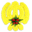 A carefully sculpted floral Khanda fills the frame: a reverent Sikh emblem rendered entirely in bright yellow chrysanthemum blooms, each tiny head packed closely to create a plush, almost textile surface. The circular chakkar, central double-edged khanda and the two crossed kirpans are all suggested by the sweep of those sunny yellow petals, their dense placement giving the symbol a slight three-dimensional lift against a clean white background. At the visual heart, where the ring meets the sword, a tight bouquet of five deep red roses is inset like a jewel, the velvety buds cradled by a collar of dark green fern fronds that add cool, feathery texture and frame the red like a small wreath. A soft shadow beneath the piece affirms it is a physical tribute-carefully freestanding or laid as a ceremonial wreath-while the stark white backdrop makes the colours sing and the craftsmanship read clearly from a distance. The scent is understated: the faint, honeyed tea-note of fresh chrysanthemum against the richer, warm perfume of newly cut roses and green fronds. As a local florist familiar with Canonbury and nearby New North Road and Canonbury Square, I imagine this used at a remembrance service, a Gurdwara commemoration, or a community cultural event in Islington-meant to offer solace, respect and a visual language of faith. Its construction shows considered layering and floral hygiene typical of bespoke memorial work from a neighbourhood workshop, created to honour tradition with a bright, uplifting palette that still carries quiet solemnity.
