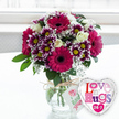 A richly textured bouquet and a whimsical heart-shaped balloon are shown together in clear, flattering daylight, as if just set down in a calm Canonbury kitchen after a surprise flower delivery. The arrangement centres around several impressive gerbera daisies in saturate magenta and fuchsia, their broad petals radiating out from dark, velvety centres that give the bouquet drama and depth. Intermingled with these are numerous small chrysanthemums with intricate two-tone petals: deep purple close to the middle, gradually lightening to sharp white at the tips, all encircling small, bright yellow centres that sparkle like tiny suns. Creamy white roses, their petals soft and layered like folded silk, are tucked evenly throughout, giving the mix an air of classic romance. Delicate sprays of baby's breath add clouds of tiny white blossoms between the more substantial flowers, balancing the strong colours and adding a hint of innocence. Lush green foliage fans out between the stems and at the base, framing the rounded bouquet and suggesting the fresh, leafy scent of a garden near New River Walk after rainfall. The flowers are gathered in a simple clear glass vase, its round shape sturdy and transparent so the straight green stems and the clean water are fully visible. A length of rustic natural twine is wrapped and tied around the vase's neck, from which a small, rectangular white tag hangs, printed with a pink rose symbol and casual green script that evokes a lovingly handmade creation. To the right, overlapping slightly with the vase, a glossy foil balloon in the shape of a heart gleams with a silver border and white centre, decorated in multi-coloured, patterned letters declaring Love n' Hugs 24-7. Set atop a pale surface in front of a softly blurred, luminous background that suggests a window in an airy Canonbury flat, the image exudes warmth, celebration, and the simple joy of sending or receiving flowers any day of the week.