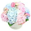 A stylish white round hat box sits alone against a completely white background, brimming with a generous, dome-shaped bouquet in soothing pastel tones that feel perfectly at home in Canonbury's calm, residential streets. The arrangement is anchored by several large, rounded clusters of light blue hydrangeas, their countless tiny petals forming soft, cloudlike shapes that lend volume and a serene, cooling presence. Tucked among these blue clouds are sumptuous pink peonies, some lush and fully open, showing off layers of ruffled, silky petals, others partially closed, as if just beginning to unfurl. Scattered delicately throughout, smaller blush spray roses appear in little groups, their buds tightly wrapped and neatly formed, bringing fine detail and a sense of gentle progression as they nestle between the larger blooms. Standing out with sculptural flair, two vivid pink calla lilies rise from the pastel mass, one to the left and one to the right, their smooth, curved trumpets adding a contemporary, almost artistic touch. At the base of the arrangement, glimpses of deep green foliage peek through, offering a quiet contrast that helps define the shape of the bouquet. The hat box itself is minimal and elegant, with a subtle ridge circling its top edge, giving the flowers a polished, gift-ready presentation that would suit a new baby welcome near Canonbury Station or a thoughtful get-well surprise on a quiet side street off Upper Street. The overall effect is one of gentle abundance, softness, and modern romance, suggesting a lovingly crafted bouquet designed to comfort, celebrate, and bring light to life's tender moments.